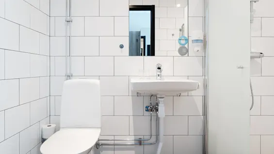 Toalett med vitt lock, stillastående, omgivet av vitkaklat badrum. Väggmonterad handfat i närheten. Spegel återspeglar rummet. Liten skylt lyder "Drink More Water" och "Go Easy On The Planet".