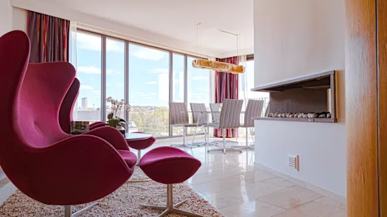 Ett modernt vardagsrum med magentafärgade fåtöljer, en matplats och en öppen spis, allt med utsikt över en naturskön vy.