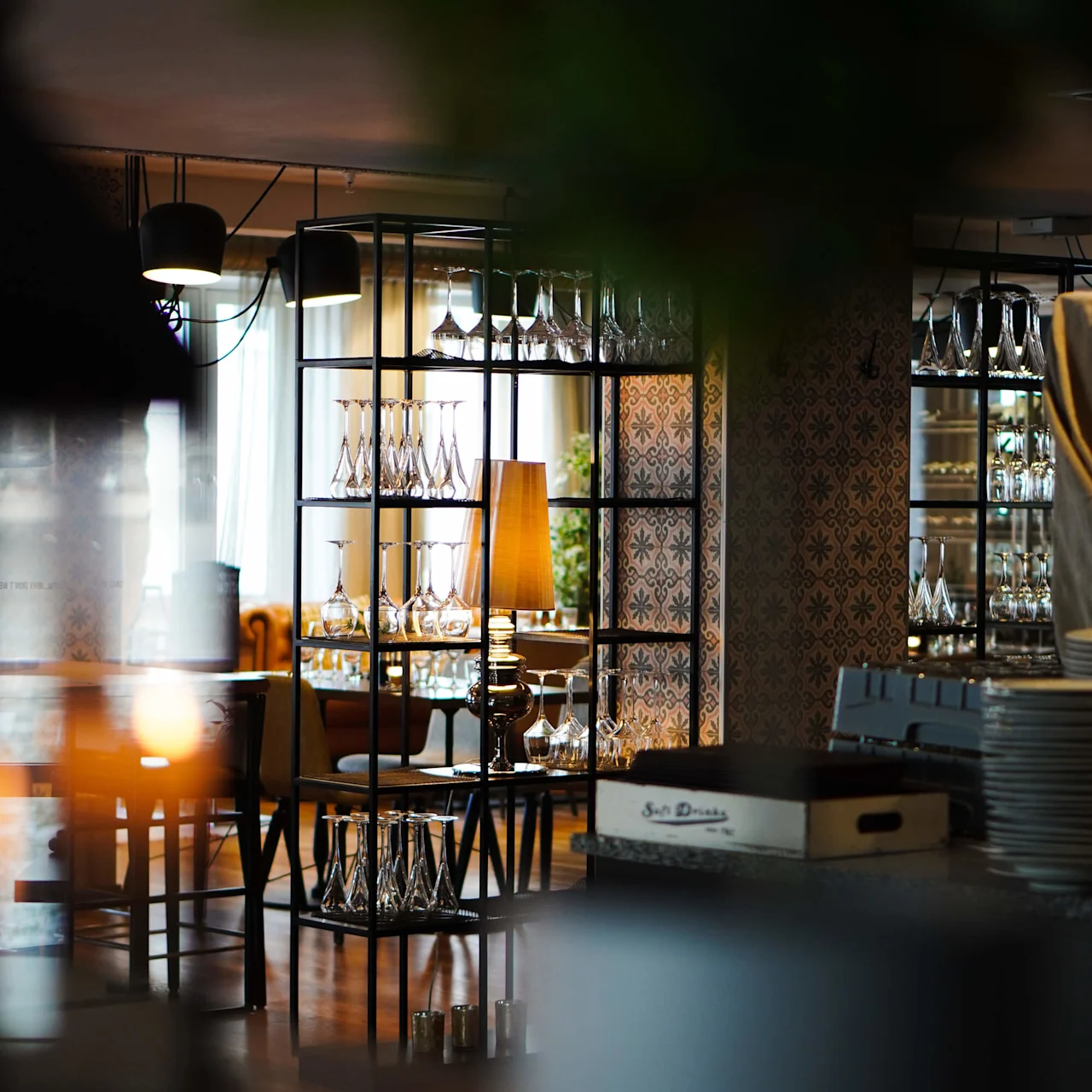 En reol viser forskellige glas i en stilfuld restaurant. Varm belysning og dekorativ tapet fremhæver den hyggelige atmosfære. En mærket kasse lyder "Soft Drinks."