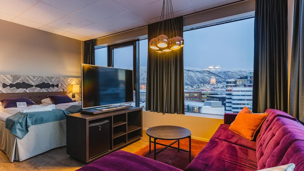 Viihtyisä hotellihuone, jossa on iso sänky, televisio ja violetti sohva, tarjoten luonnonkauniin näkymän lumisille vuorille ja kaupunkiin.