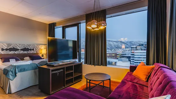 Et koselig hotellrom med en stor seng, en TV og en lilla sofa, med en naturskjønn utsikt over snødekte fjell og en by.