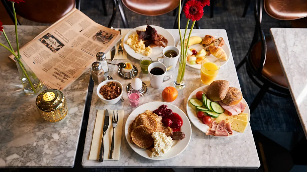 Frukostbord med pannkakor, ägg, bacon, frukt, ost, skinka, croissanter, kaffe och juice. Röda blommor i en vas och en öppen tidning kompletterar miljön.