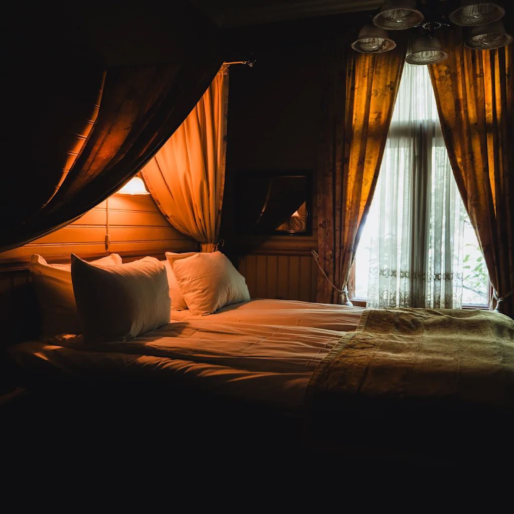 A cozy room at Dalen Hotel, warmly lit, with a plush bed next to a window draped in rich curtains, casting a serene ambiance.