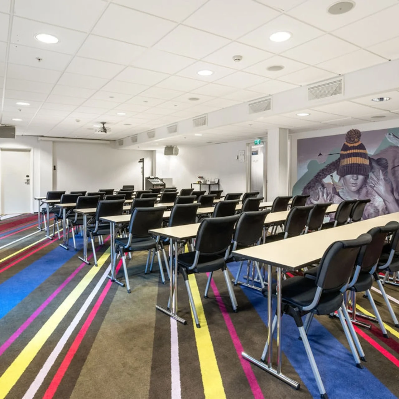 Rows of black chairs face a projection screen in a colorful carpeted conference room. A mural of a person beside a reindeer adorns the wall, enhancing the modern decor.