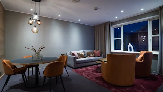 A cozy living room features a gray sofa and orange armchairs on a red rug, under dim, decorative lights, beside a window with beige curtains.