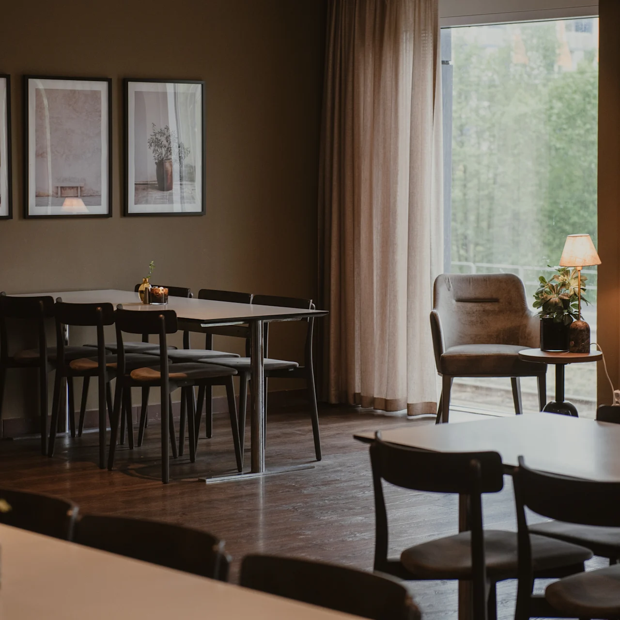 Several wooden tables and chairs are arranged in a warmly lit room. Framed artwork hangs on the wall, and a window shows green trees outside. A lamp and small plants decorate tables.