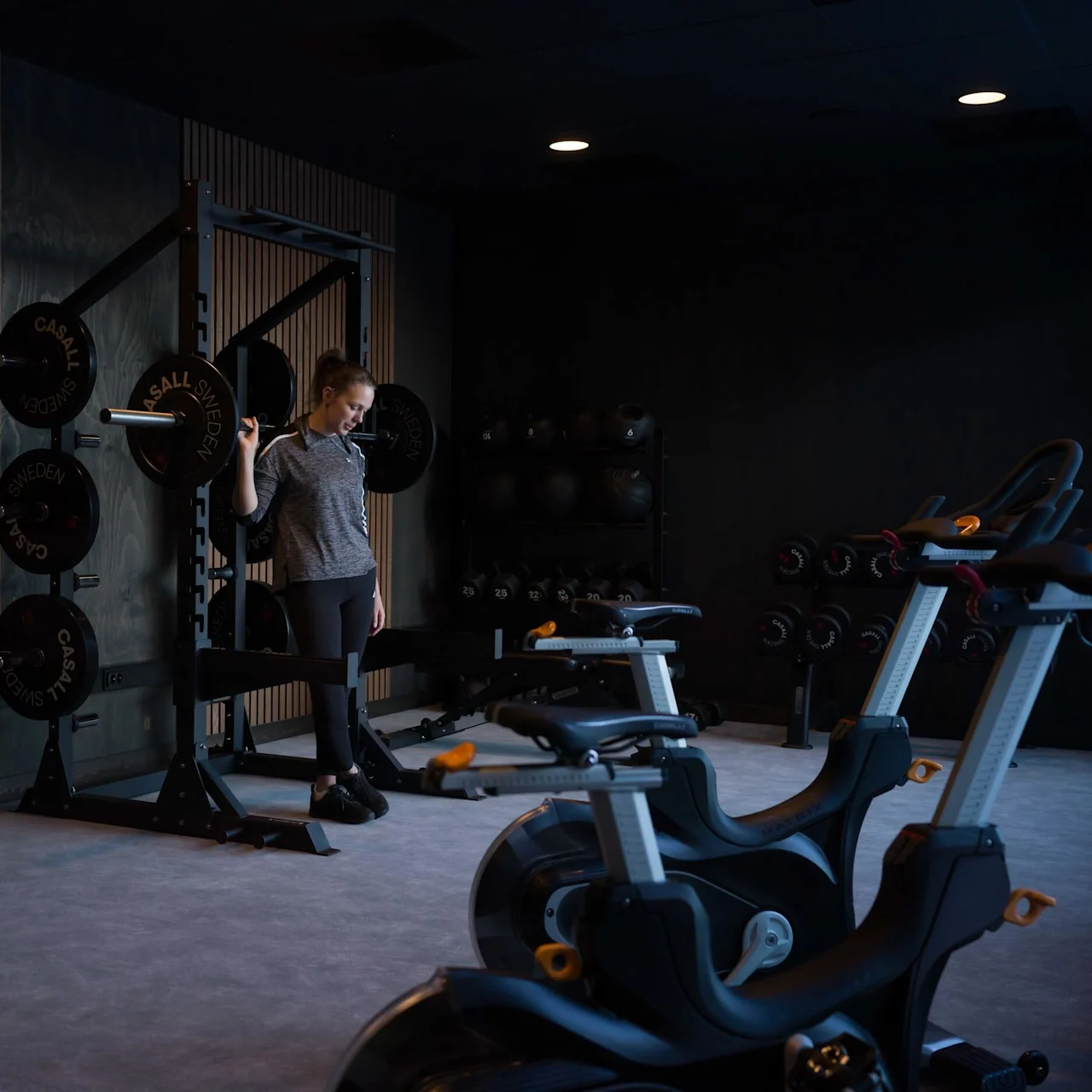 A person holds a barbell in a gym setting, surrounded by exercise bikes, weights, and kettlebells. The walls are wooden, and the room is dimly lit. Text on weights: "CASALL SWEDEN."