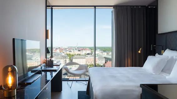 Et romslig hotellrom har en pent oppredd seng, en flatskjerm-TV og et stort vindu med panoramautsikt over byen. En polstret stol står nær vinduet.