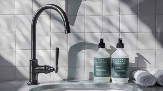 Tap with dark finish stands idle next to two soap bottles labeled "SKANDINAVISK SKOG" on a tiled bathroom counter. Rolled towels lie nearby.