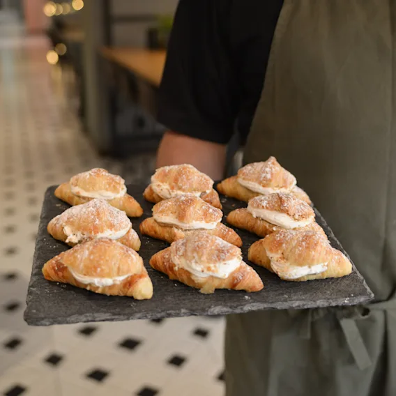 Nine sugar-dusted pastries with cream on a black tray are carried by a person wearing a green apron in a tile-floored cafe.