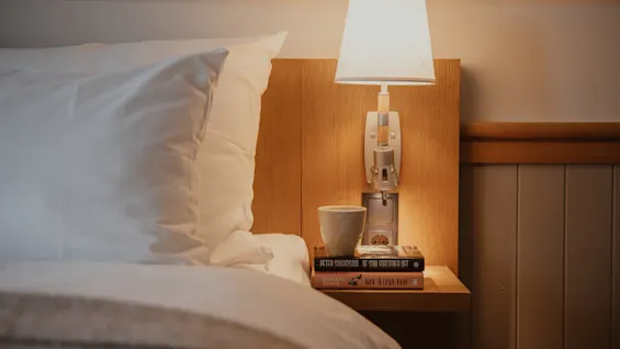A cozy bedside scene with a warm lamp, a cup, and books on a nightstand next to a bed with white linens.