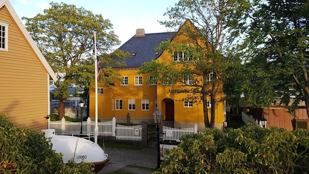 A vibrant yellow building with a dark roof, nestled among lush green trees and a white picket fence, under a clear sky.