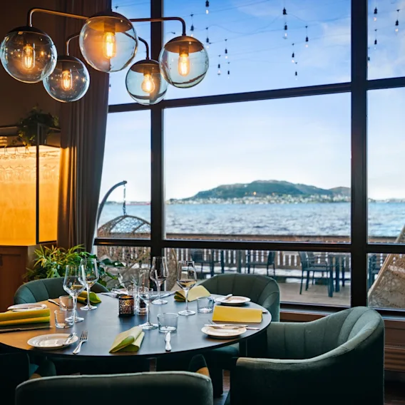Dekket rundt spisebord i en moderne restaurant med grønne fløyelsstoler og store vinduer med utsikt mot fjorden.