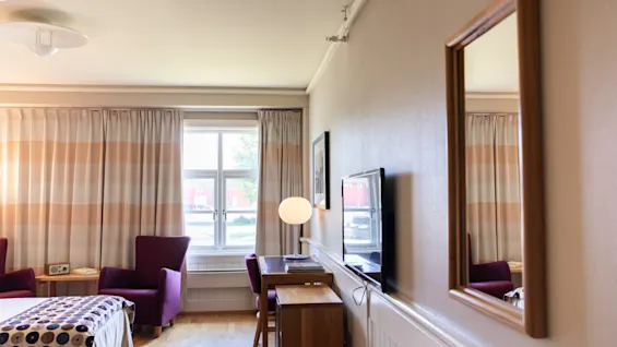 Et hotelværelse har en seng med prikket betræk, to lilla stole, et skrivebord med lampe og et vægmonteret TV. Gardiner er trukket for ved siden af et stort vindue.