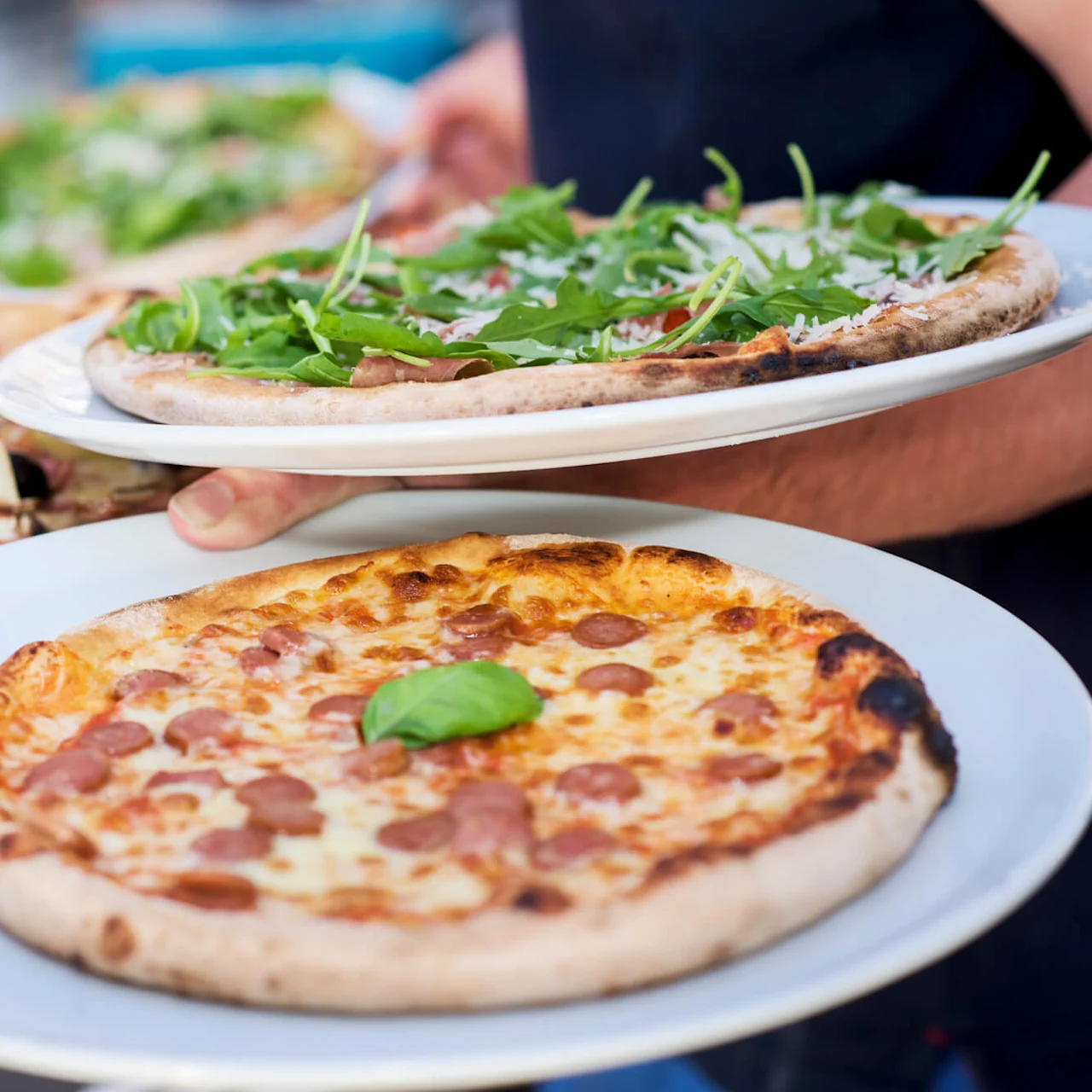 Fire pizzaer på hvite tallerkener serveres, med toppinger som basilikum, ruccola, sopp og charkuteri. Bakgrunnen antyder en uteplass.