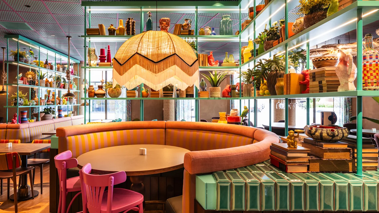 En buet bod med et rundt bord og farverige stole omgivet af eklektisk indretning og forskellige genstande på hylder, der skaber en livlig, hyggelig caféatmosfære.
