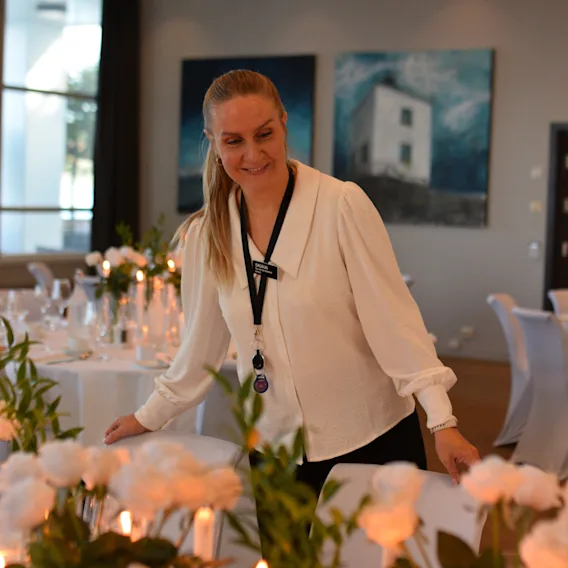 En smilende kvinne i hvit bluse som retter på en stol i et festlokale pyntet til bryllup med hvite blomster.