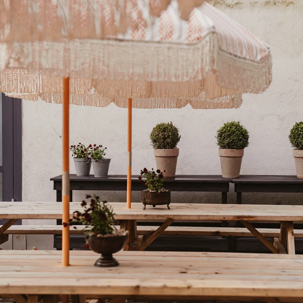 Wooden picnic tables with decorative potted plants sit beneath fringed umbrellas in a courtyard with potted shrubs on a bench against a textured wall. A watering can is nearby.