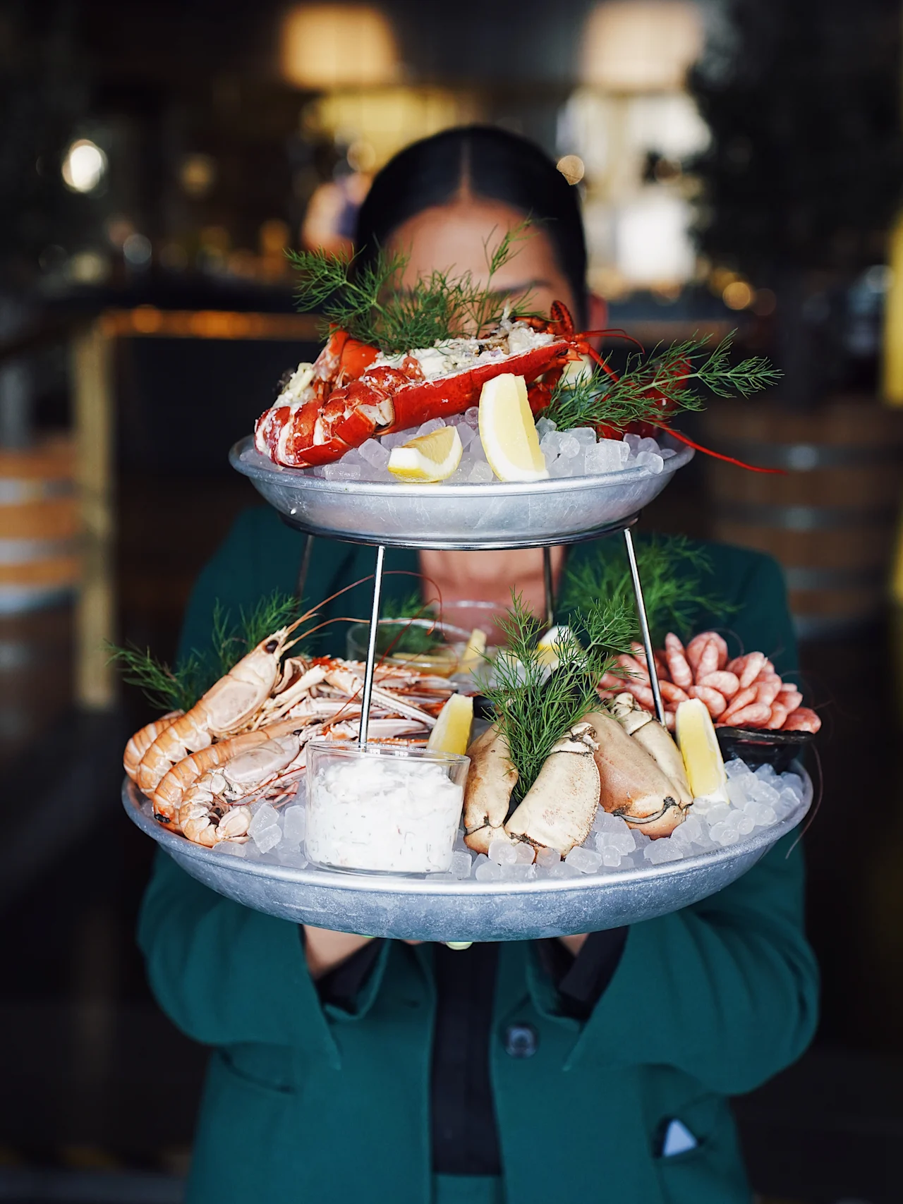 Tiered seafood platter with lobster, prawns, and clams resting on ice, garnished with lemon wedges and dill, being held by a person in a restaurant setting.