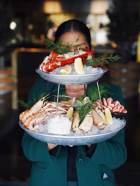 Tiered seafood platter with lobster, prawns, and clams resting on ice, garnished with lemon wedges and dill, being held by a person in a restaurant setting.