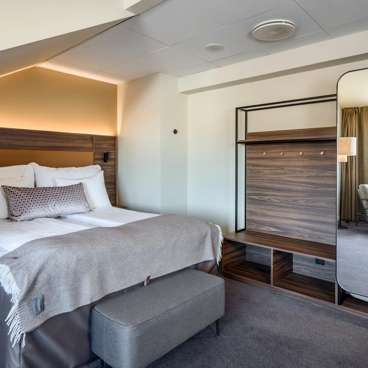 Bed adorned with pillows and a blanket sits in a modern room. A large mirror and wardrobe frame the space. A window admits natural light onto a chair.