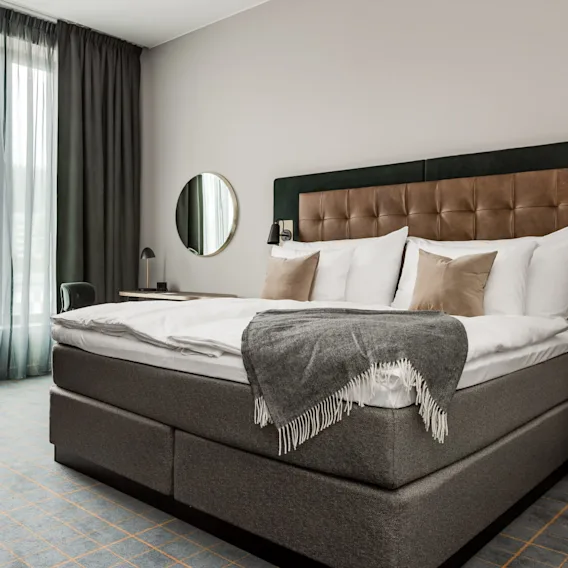 A neatly made bed with pillows and a grey blanket is in a hotel room, accompanied by a sofa, side tables, and a large window with curtains.