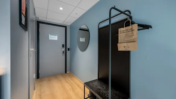 En hotellromsinngang med dør, speil og en kleshenger. En papirpose på hengeren sier 'DU OG DIN T-SKJORTE KAN GJØRE EN FORSKJELL'.