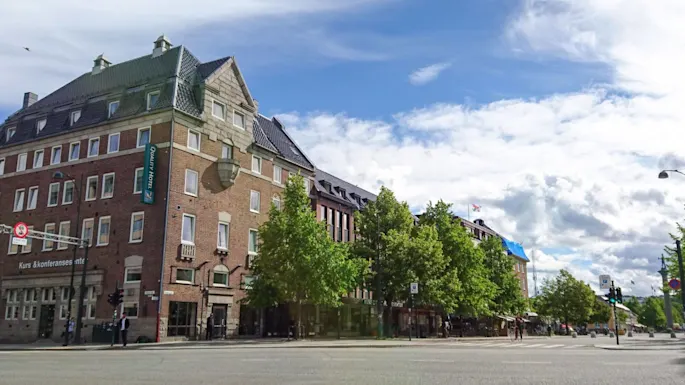 Tiilitalohotelli, jolla on tiilikatto, seisoo kadunkulmassa. Sininen taivas, pilviä, ja puita tien varrella. Kylttejä, joissa tekstit "Quality Hotel" ja "Kurs & konferansesenter."