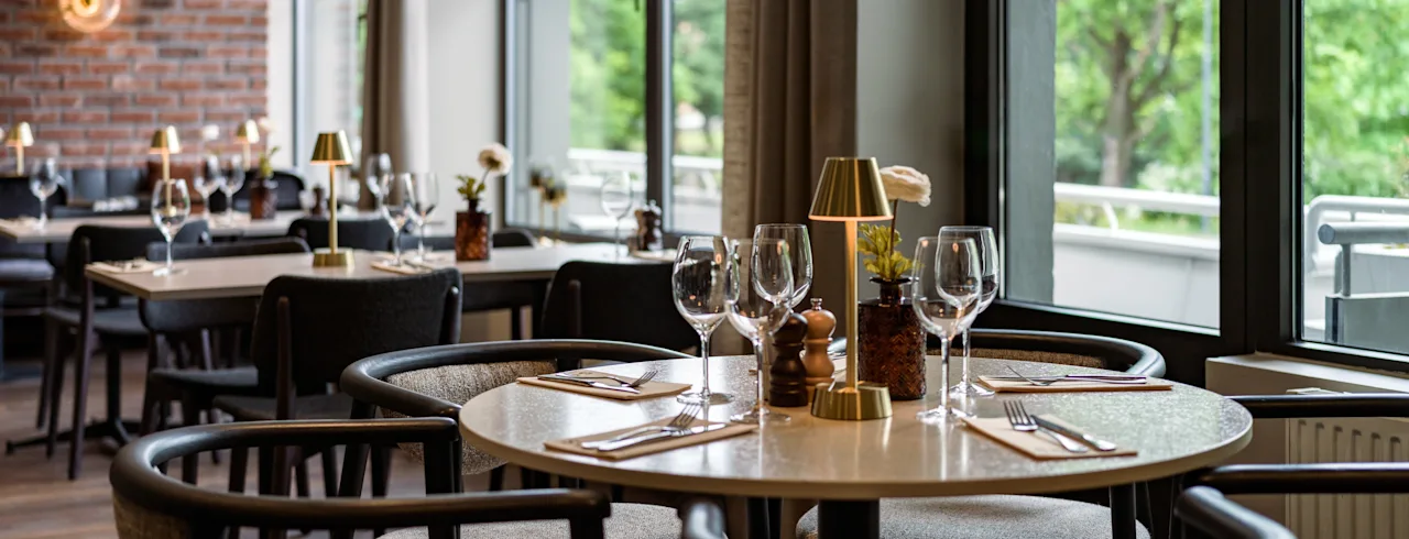 Modernt matbord med vinglas, bestick och en liten lampa i en restaurang med tegelväggar och stora fönster som vetter mot gröna träd.