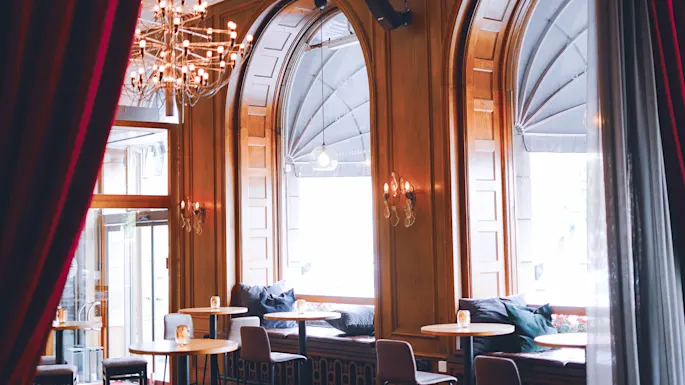 Elegant caféindretning med lysekrone, bueformede vinduer og hyggeligt siddeområde i naturligt lys.