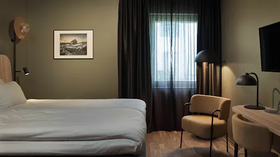 Ett mysigt hotellrum med en säng, ett skrivbord och ett fönster med mörka gardiner, vilket skapar en bekväm och inbjudande atmosfär.