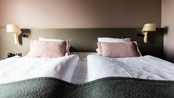To nøye oppredde enkeltsenger med rosa puter, flankert av vegglamper, i et rent, moderne soverom.