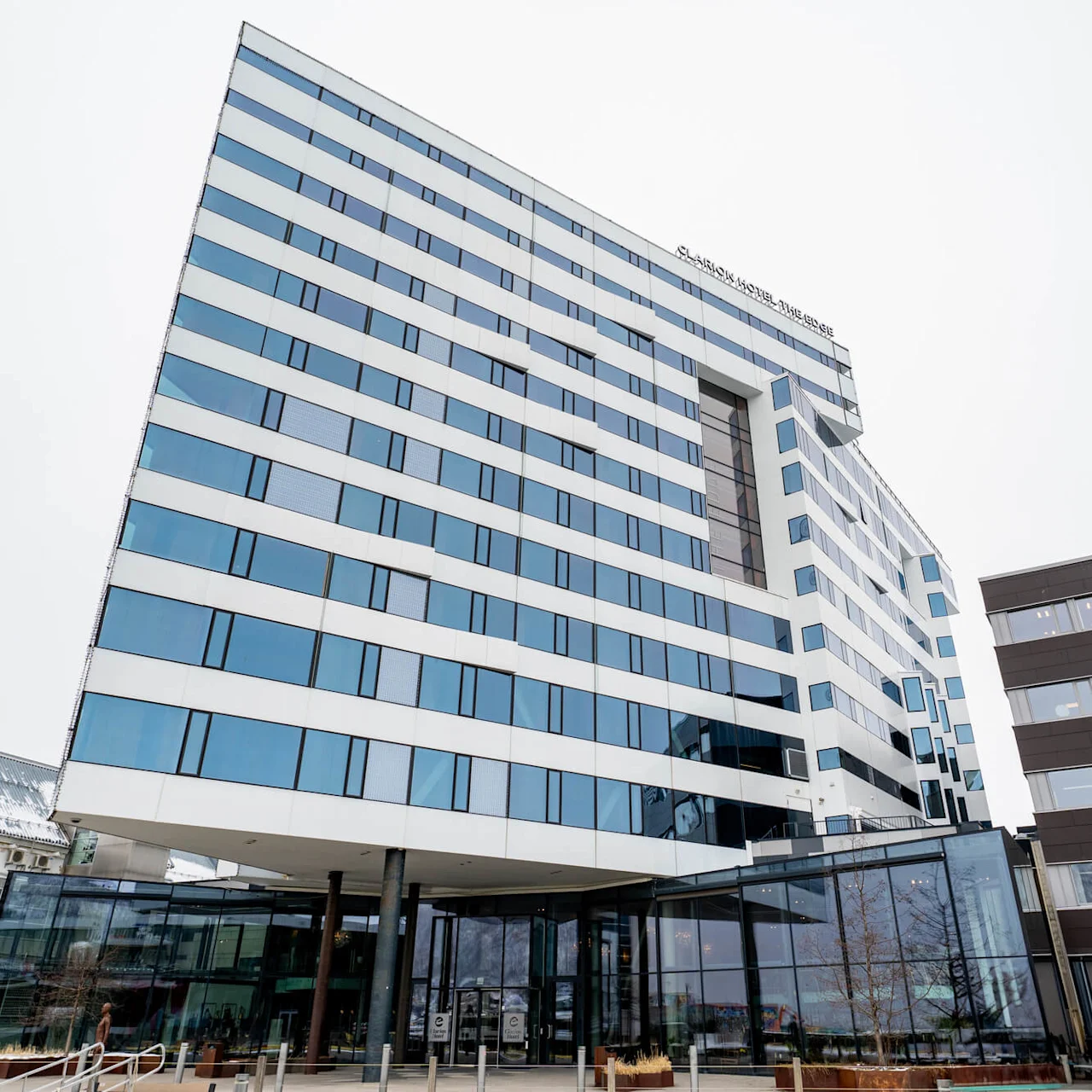 En moderne bygning i flere etager med store glasvinduer er beliggende i et byområde med yderligere strukturer i nærheden. Tekst på bygningen lyder: "CLARION HOTEL THE EDGE".