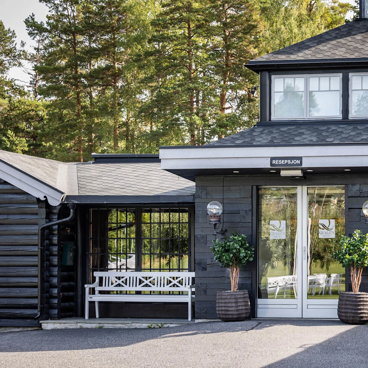 Inngang til en hotellresepsjon med glassdører, flankert av potteplanter og benker; omgitt av skogsområde. Tekst på døren: "QUALITY HOTEL ENTRY", "RESEPSJON".