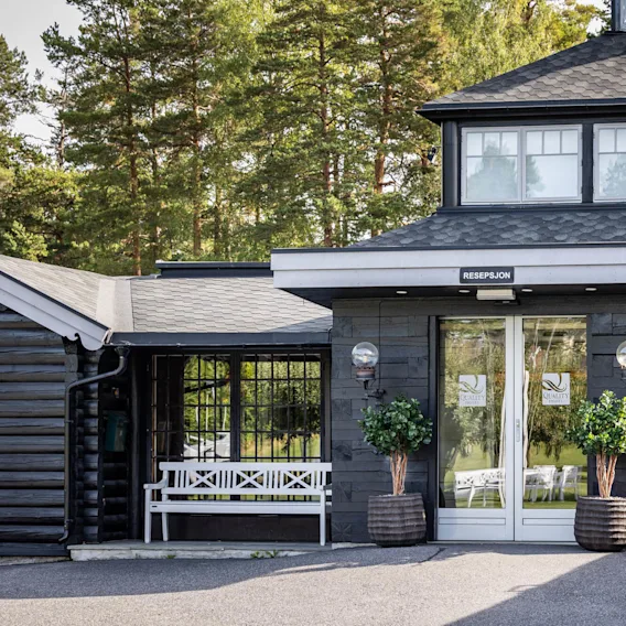 Inngang til en hotellresepsjon med glassdører, flankert av potteplanter og benker; omgitt av skogsområde. Tekst på døren: "QUALITY HOTEL ENTRY", "RESEPSJON".