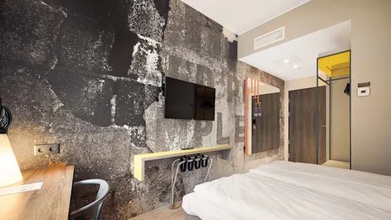 A hotel room features a bed with white linens, a wall-mounted TV, and a rustic wall texture. A desk with a chair sits nearby. Text on wall: "OPLE".