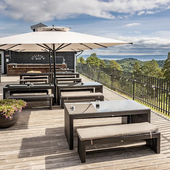 Utendørs terrasse med trebord og benker under en stor parasoll med utsikt over et vakkert landskap