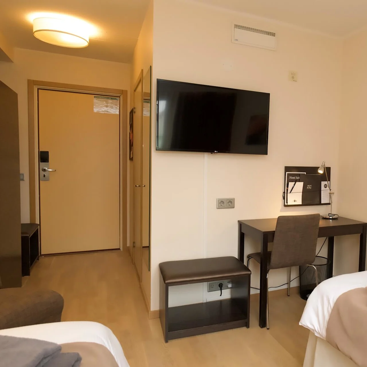 Et hotelværelse med en redt seng, sammenfoldede håndklæder og en fjernbetjening ovenpå, et vægmonteret TV og et skrivebord nedenunder. En lukket dør fører udenfor.