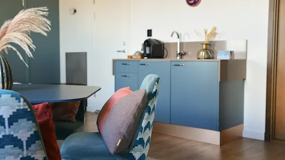 Blå stole med mønstret polstring omgiver et spisebord, dekoreret med farverige puder. I baggrunden er der et kompakt køkken med vask, kaffemaskine og dekorative genstande på bordet.