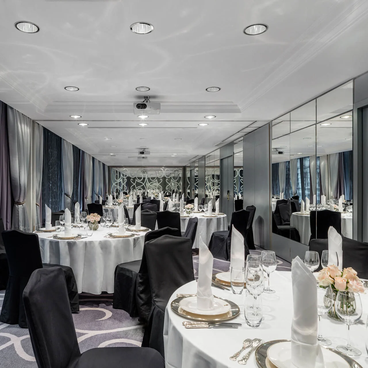 Elegant dining tables set with white linens and black chair covers stand in a luxurious banquet room, with mirrored walls and large windows draped with sheer curtains.