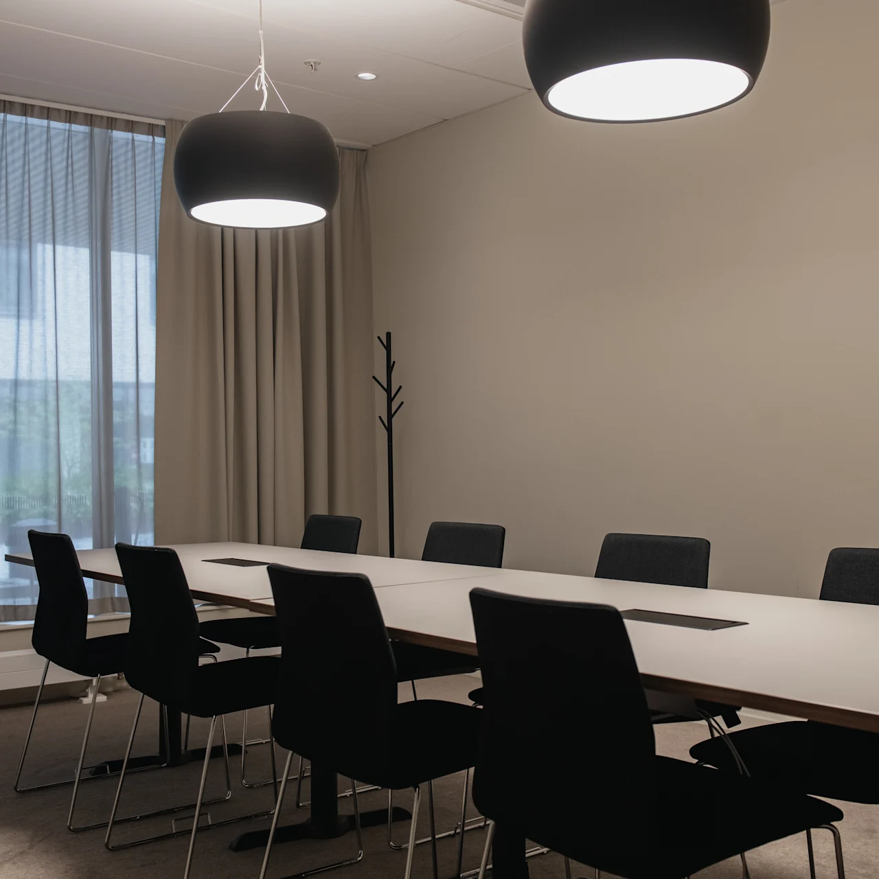 A long conference table surrounded by eight black chairs rests under two ceiling lamps, in a softly lit meeting room with a large window covered by sheer curtains.