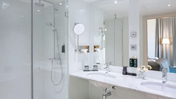 Brusebad lukket i glas ved siden af marmorbordplade med dobbeltvaske i et moderne, lyst badeværelse. Tilstødende værelse synligt med seng og gulvlampe.