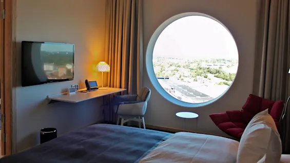 A round window overlooks a sunny landscape in a modern hotel room, featuring a bed, wall-mounted TV, desk with a lamp, and a red chair creating a cozy atmosphere.