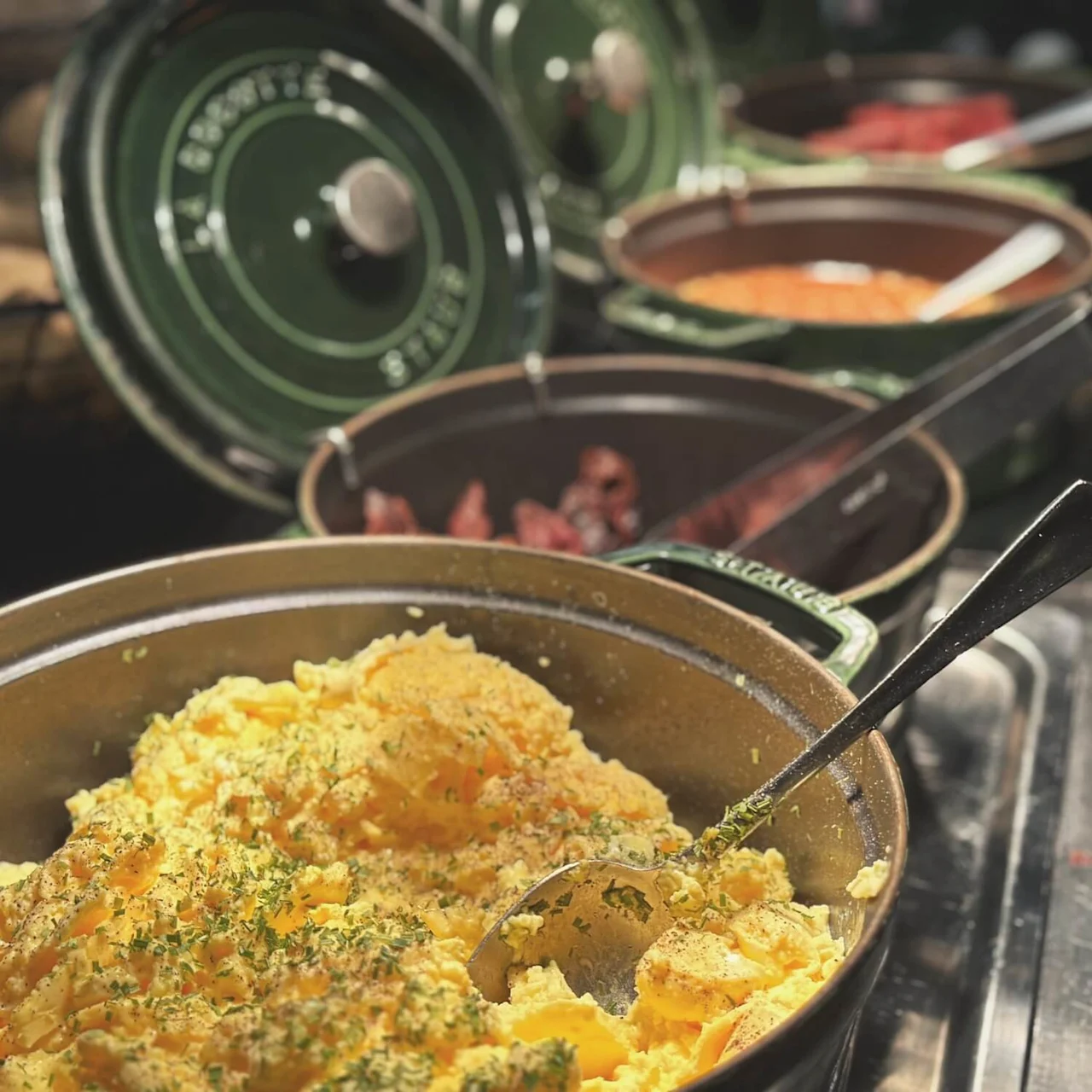 A close-up of a breakfast buffet featuring a pot of steaming scrambled eggs garnished with herbs. In the background, several pots contain various breakfast items, including sausages and baked beans.