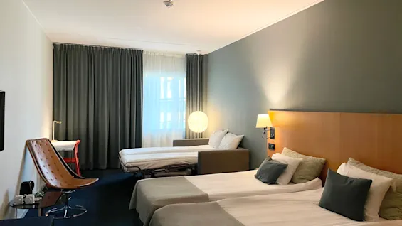 To pent re oppredde senger med luksuriøse puter og tepper er i et hotellrom. Et skrivebord og stol står ved siden av nær et gardinert vindu, og skaper en koselig atmosfære.