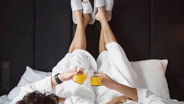 Två personer i badrockar och tofflor kopplar av i sängen och skålar med apelsinjuice. En mysig och glad stund av avkoppling.