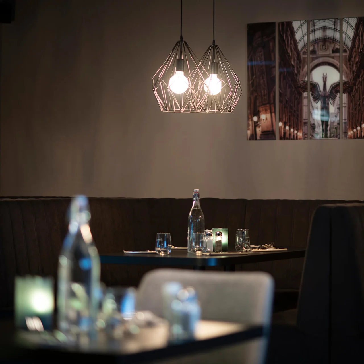 Hänglampor lyser upp mörka sammetssoffor med bord dukade med vattenflaskor och glas i en svagt upplyst restaurang, med ett stort väggfoto av en arkitektonisk scen.