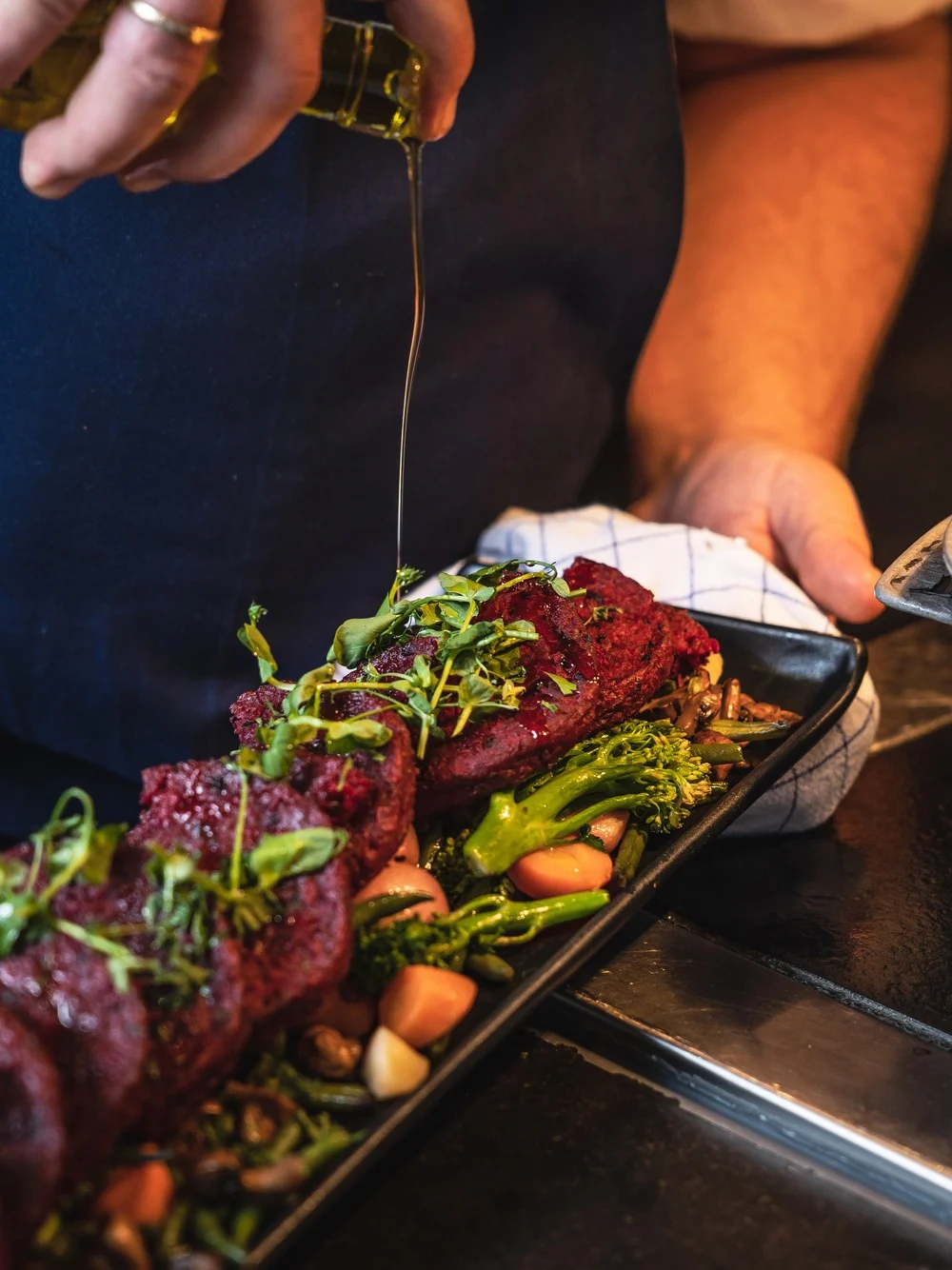 Kokker hell olje over skivet kjøtt med urter og grønnsaker på et mørkt fat, ved siden av en gryte med kokte kyllingstykker i kjøkkenmiljø.