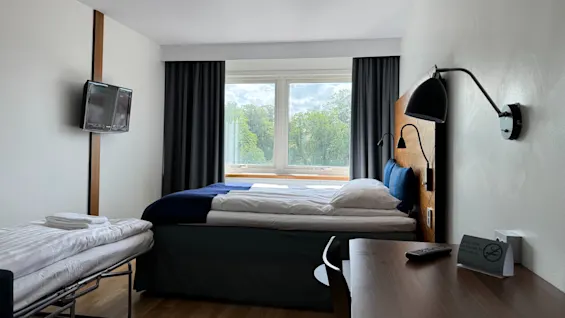 A neatly made hotel bed stands against a window with drawn curtains, flanked by a wall-mounted lamp. Nearby, a wooden desk and a folded spare bed. 