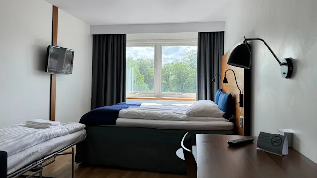 Sänky hotellihuoneessa on ikkunaa vasten verhojen edessä, vieressään seinävalaisin. Lähellä on puinen pöytä ja taitettava varavuode.